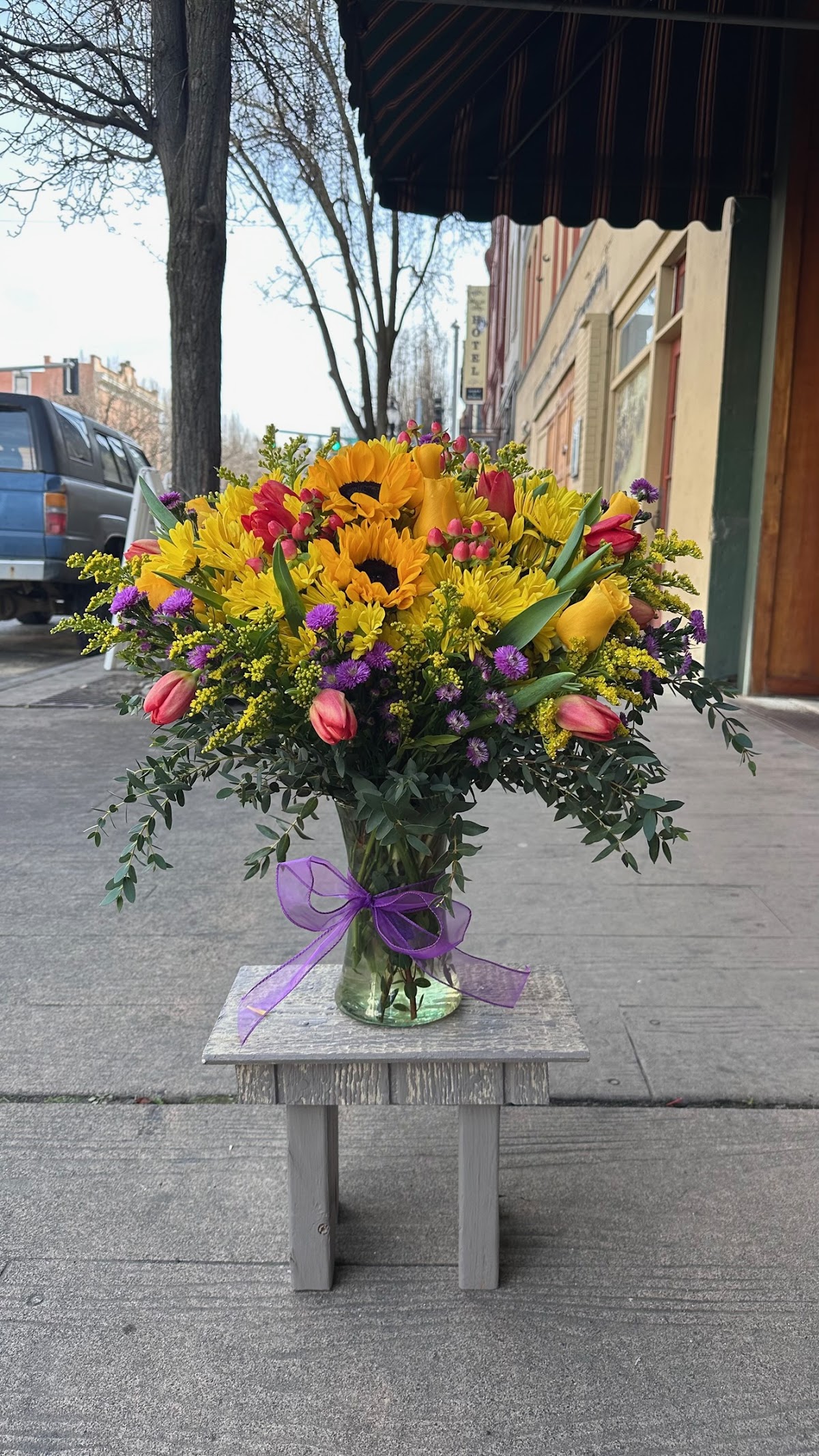 A bright hand-arranged bouquet of sunflowers, tulips, and daisies in a vase with a purple bow