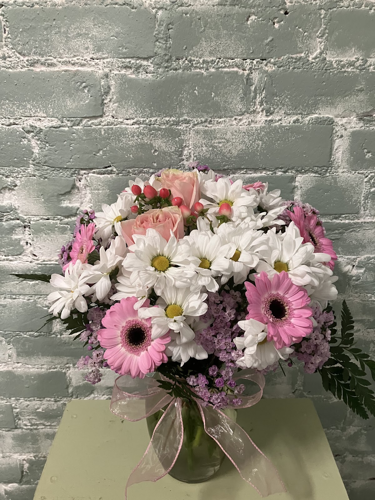 A soft pink and white arrangement of gerbera and daisies on the sage-green brick wall